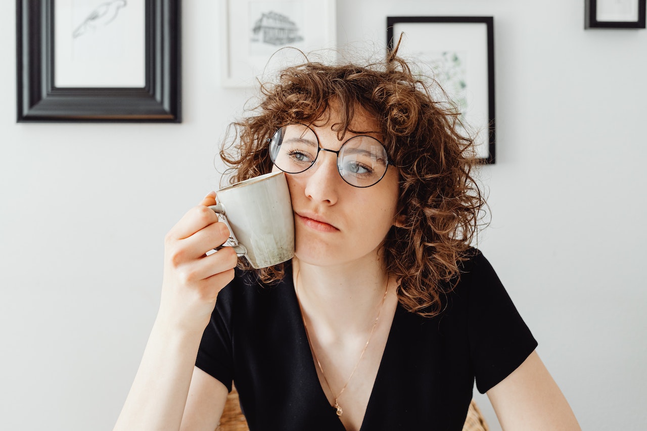 A Caucasian women wearing glasses is overthinking things while holding a cup to her left cheek of her face while sitting in an office.